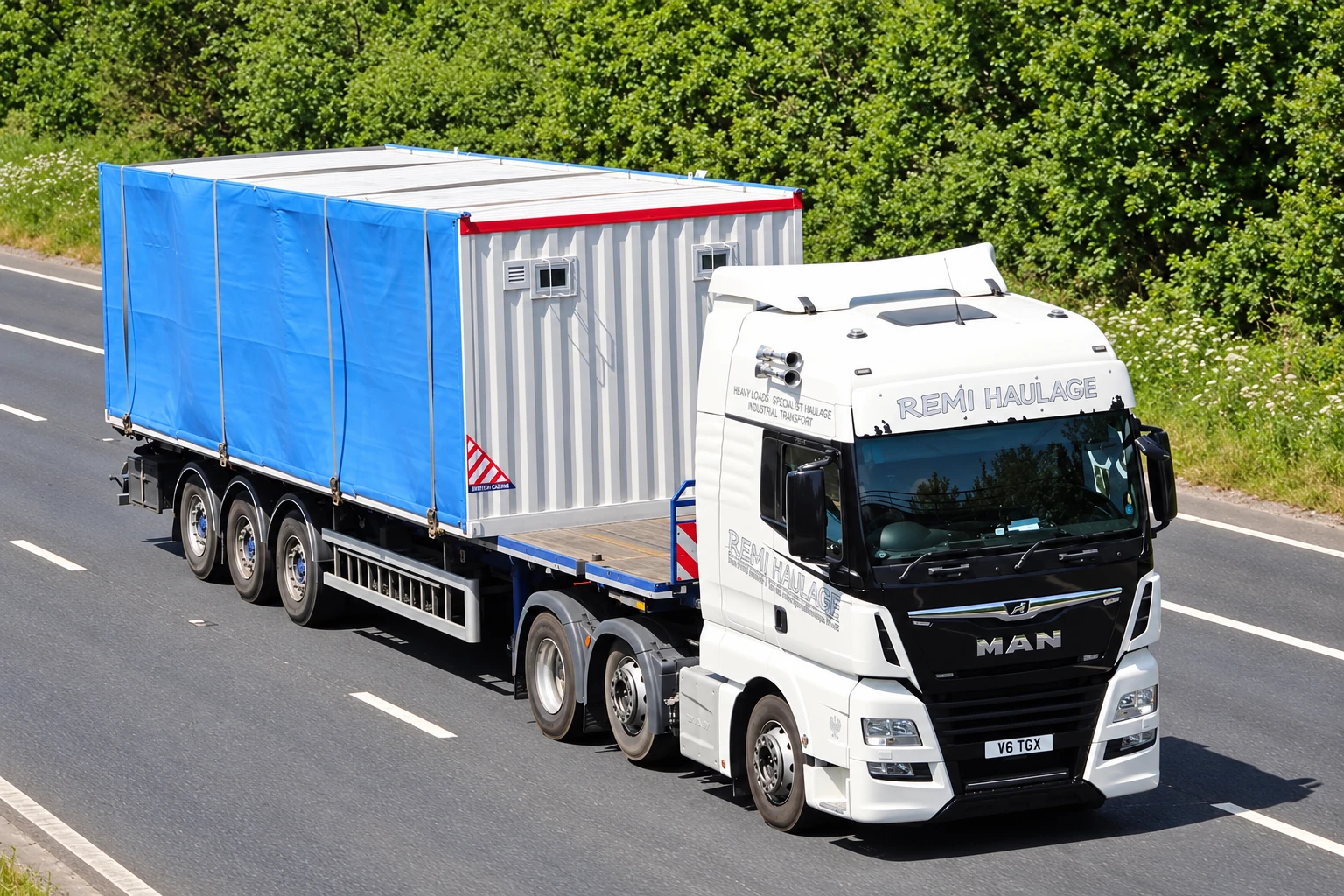 White Remi Haulage MAN truck transporting a blue-wrapped modular cabin on a UK motorway